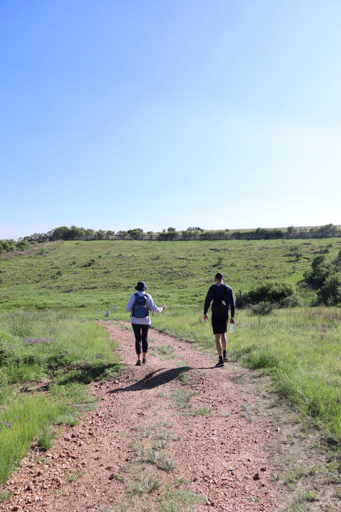 Two individuals walking along a gravel path with a scenic backdrop of green grass landscape